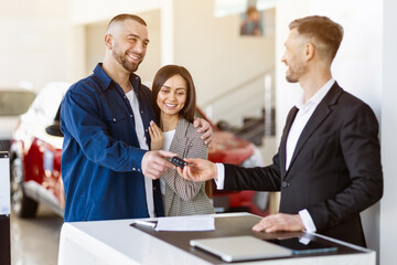 Smiling couple receives car key from dealer at dealership office. Young family buying auto together, joyful moment of purchase and ownership in bright and modern environment