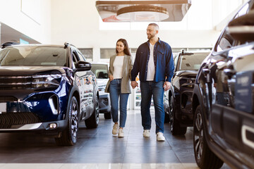 Young couple holding hands while walking through modern car showroom. Joyful and relaxed atmosphere...
