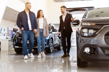 Car salesman showing vehicle details to cheerful couple inside showroom. Smiling pair stands close together, enjoying the car shopping experience, full length shot