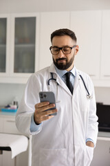 Handsome male Hispanic doctor in medical coat and glasses browsing Internet on mobile phone on work...