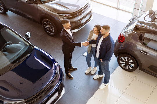 Dealer shaking hands with young couple in car showroom from aerial view. Symbolic moment of trust and closure in vehicle purchase decision, above shot