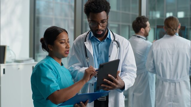 Doctors cooperation teamwork Indian Arabian woman nurse therapist asking advice African American man doctor doc pediatrician therapist practitioner discussing holding digital tablet device at clinic - Powered by Adobe
