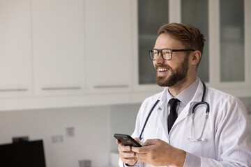 Positive young medical practitioner man in eyewear and lab coat typing on smartphone in work...