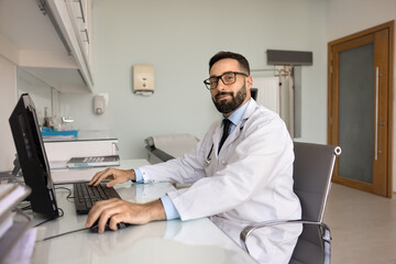 Handsome Hispanic male physician in glasses and lab coat working at computer in clinic office,...