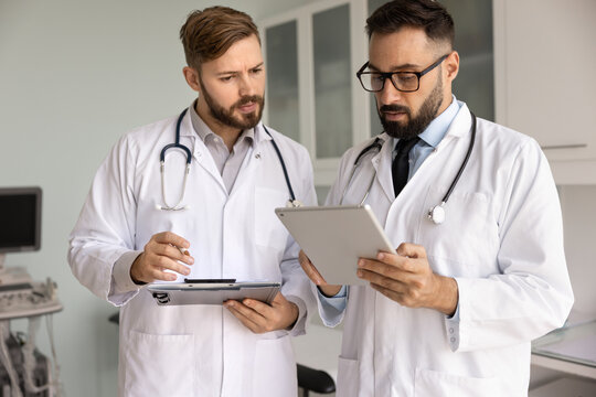 Two serious doctors in lab coats discussing medical case, examination electronic results, standing in hospital office together, using tablet computer, digital pad, checking records