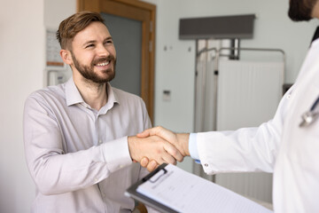 Cheerful positive young Caucasian male patient giving handshake to doctor in lab coat, thanking...