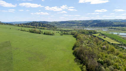 Obraz premium Aerial view of lush green hills and valleys under a bright blue sky with scattered clouds, showcasing the beauty of nature in a tranquil landscape. Ideal for outdoor themes.