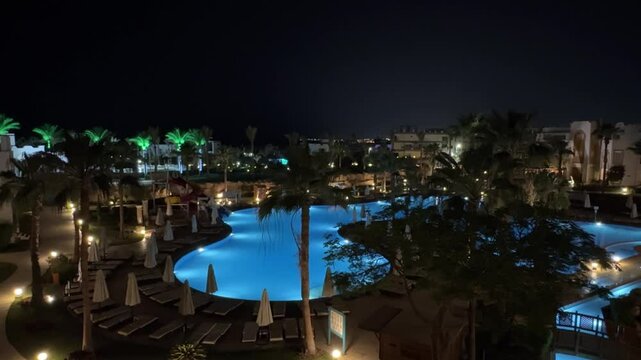 Nighttime view of a luxurious resort pool illuminated by soft lights, surrounded by palm trees and lounge chairs, creating a serene atmosphere for relaxation and leisure activities in a tropical parad