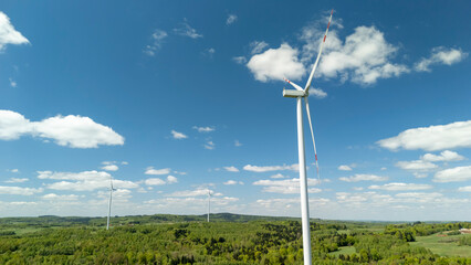 Wind turbines stand tall against a bright blue sky, surrounded by lush green hills and trees,...