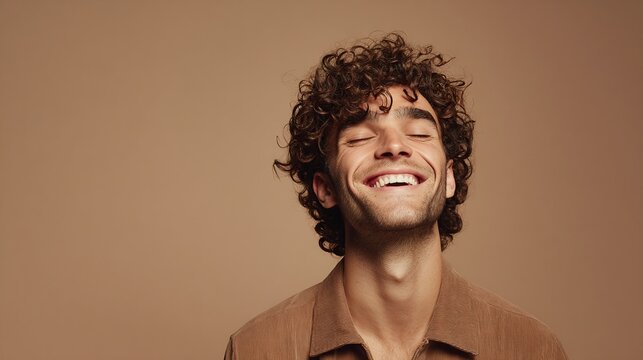 Radiant joy: A young man with curly hair smiles serenely against a warm background, eyes closed in contentment. Expression of inner peace.
