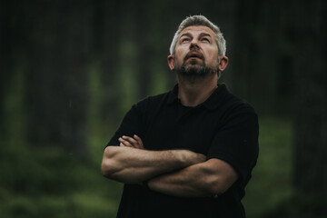 A man with gray hair and a beard stands with his arms crossed, looking up into a shady forest. He is wearing a black shirt, and his mood is contemplative and calm.