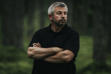 A man with gray hair and a beard stands with his arms crossed, looking up into a shady forest. He is wearing a black shirt, and his mood is contemplative and calm.