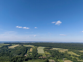 Aerial view of lush green fields and forests under a clear blue sky. The landscape features varied agricultural lands and a rural village, showcasing natural beauty.