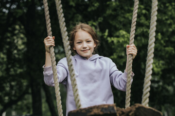 A cheerful girl in a lavender hoodie holds the ropes of a wooden swing in a park surrounded by deciduous trees and smiles directly at the camera. Natural daylight and greenery in the background.