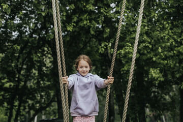 A cheerful girl in a lavender hoodie holds the ropes of a wooden swing in a park surrounded by deciduous trees and smiles directly at the camera. Natural daylight and greenery in the background.