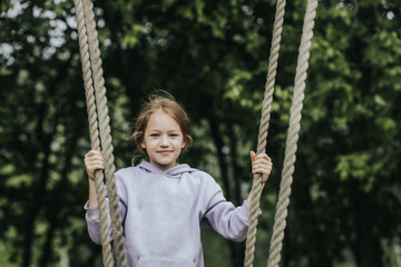 A cheerful girl in a lavender hoodie holds the ropes of a wooden swing in a park surrounded by deciduous trees and smiles directly at the camera. Natural daylight and greenery in the background.