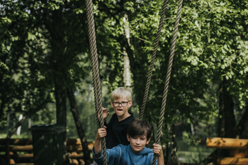 Two boys sit on a rustic wooden swing suspended by ropes in a lush green park surrounded by trees and wooden benches, enjoying a peaceful moment outdoors.