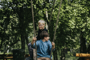 Two boys sit on a rustic wooden swing suspended by ropes in a lush green park surrounded by trees and wooden benches, enjoying a peaceful moment outdoors.