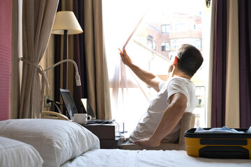 A digital nomad man with a laptop computer, waiting for an online meeting, opens the curtains to...