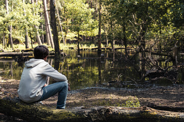 A boy rests on a tree trunk, gazing at the tranquil pond in the forest. The warm sunlight filters...