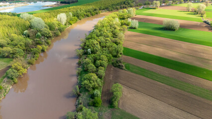 Aerial view of a river winding through lush green trees and vibrant agricultural fields in springtime, showcasing the beauty of nature and rural landscapes.