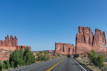Drivers perspective over the road approaching The Courthouse Towers landmark geological formation and Three Gossips formation against a blue sky in Arches National Park, Moab, UT, USA