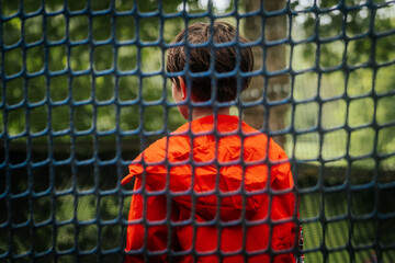 A child in a vivid orange jacket stands with his back to the camera, framed by blue netting in an outdoor wooded setting.