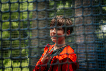 A cheerful boy in an orange jacket poses with a peace sign behind a blue safety net outdoors, in a playful atmosphere, surrounded by greenery.