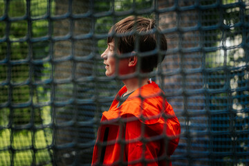 A young boy in a bright orange jacket smiles in profile behind blue netting outdoors, with trees...