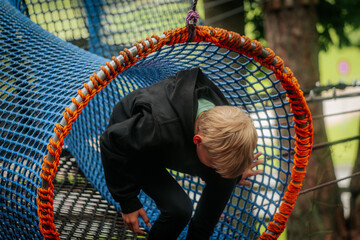 A small child in a black hoodie moves through a colorful rope tunnel made of blue and orange netting in a forest adventure park.