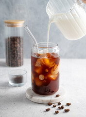 Milk pouring from a milk jug into a glass of iced coffee on a marble board on a light background