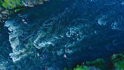 Aerial view of a kayak boat rushing along a turbulent river between stone rocks at high speed.