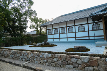 Minimalist Japanese Zen rock garden, or karesansui, sits beside a traditional white-walled building, its raked gravel and mossy stones creating a tranquil space for meditation and reflection in Kyoto.