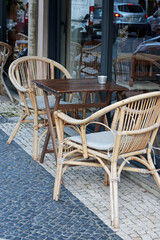 Outdoor cafe scene with wicker chairs and wooden table on tiled pavement. Cozy corner for coffee breaks and casual dining, perfect for travel-themed prints, hospitality projects, or lifestyle designs.