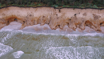 Aerial view of ocean waves beating against a rocky shore on a wooded tropical seaboard.