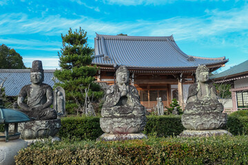 Ancient, weathered stone statues of Buddha and Bodhisattvas sit serenely in a manicured garden before a traditional temple hall at the Ninna-ji World Heritage site in Kyoto, Japan, on a bright day.