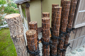 Traditional Japanese fence, constructed from bundles of twigs bound with copper wire and black rope, stands beside wooden building, showcasing intricate craftsmanship and natural materials in a garden