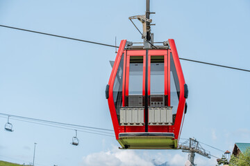 The red cab on the cable car stands against the backdrop of a summer forest and mountain peaks.