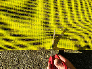Close-up of a person cutting yellow fiberglass facade mesh with red-handled scissors on a rough...