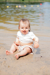 adorable baby enjoys playful moment on a sunny day at the beach, with sandy hands and feet, wearing a white shirt, against a backdrop of gentle lake waves and colorful buoys. gentle, soft, vibrant