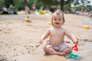joyful toddler playing with sand and toys outdoors on a sunny day in a park with families and trees in the background, recreation, summertime, fresh, breezy, vacation, holiday, delight, innocent
