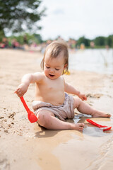 adorable toddler enjoying playful beach time with toys by the water on a sunny day, surrounded by a scenic, vibrant outdoor setting with sand, trees, and distant figures. sunshine, sandcastle, bliss