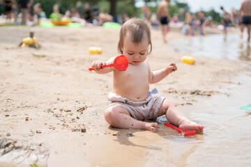adorable baby enjoys playful moment with red shovel on busy beach, surrounded by sand, water, and a vibrant summer atmosphere with families and children in the background, joyful, vibrancy
