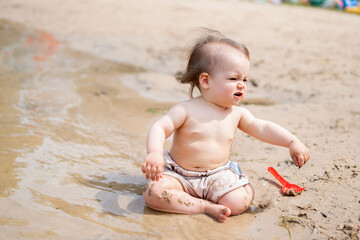 a playful toddler sits on a sandy beach, joyfully exploring the wet sand with a red toy shovel, capturing a moment of carefree summer fun and early childhood discovery. moment, spontaneous
