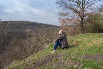 Man sitting on hill overlooking a valley enjoying the serene landscape on a cloudy day in early...