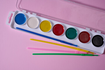 A set of watercolor paints in a plastic case with multi-colored brushes is located on a pink...
