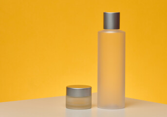 Two matte transparent cosmetic bottles with silver caps, a tall cylindrical one and a short round one, yellow background