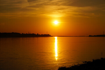 Naklejka premium an orange evening sunset over the Kostroma River with the reflection of the sun and cirrus clouds in the water, a view from the shore in the city of Kostroma, the opposite shore with trees is visible