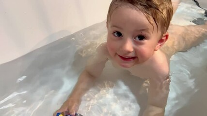 Two-year-old little cute child boy bathing in water in a white bathtub, playing with toys