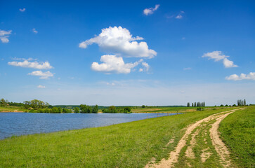 Rural landscape with dirt road beside a tranquil lake under blue sky
